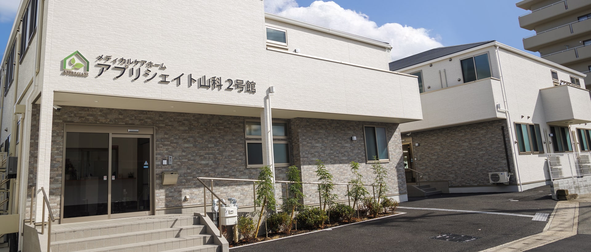 介護施設の建物外観。白とグレー基調の2階建て施設で、玄関前に植栽がある。